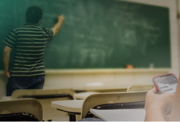 Student playing on phone while a teacher is writing on a blackboard.